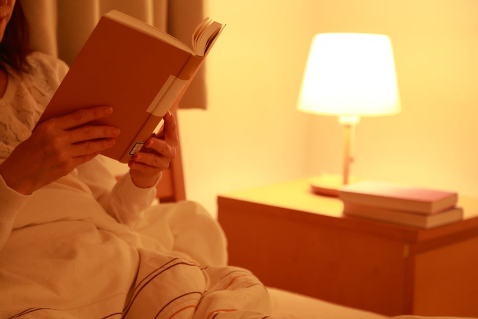 ベッドサイドに置いたスタンドライトの光で読書をする女性
