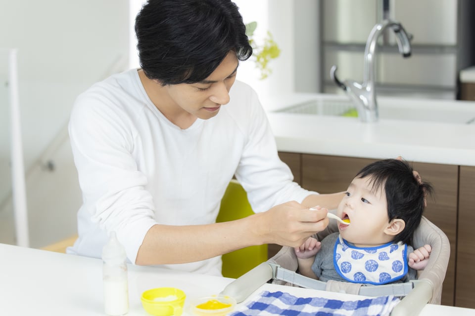 卓上設置ベビーチェアに乳児を座らせ離乳食を食べさせる父親